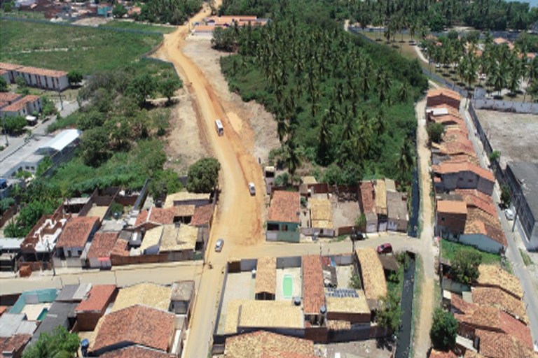 Esta imagem aérea exibe uma perspectiva elevada da Comunidade do Estaleiro, em Barra dos Coqueiros (SE), destacando o contraste entre a urbanização e as intervenções de infraestrutura. No centro, uma estrada de terra batida em tons alaranjados corta o cenário, onde se vê um ônibus e alguns veículos pequenos em movimento. À esquerda e na parte inferior, há um aglomerado de casas com telhados de barro e algumas construções com pequenas piscinas nos quintais, enquanto o lado direito é dominado por uma extensa área verde com muitos coqueiros, típica da região litorânea. O registro, creditado ao MPF, documenta o impacto de obras e desapropriações na localidade, evidenciando a proximidade entre a zona residencial e a área de preservação ou cultivo.