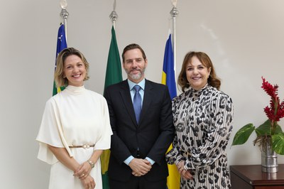 Fotografia em plano médio mostra três pessoas sorrindo enfileiradas lado a lado em um ambiente institucional, com três bandeiras oficiais ao fundo. À esquerda, uma mulher de cabelo loiro curto veste um vestido branco com cinto fino e mangas fluídas; ao centro, um homem de barba curta e terno escuro usa camisa azul-clara e gravata azul; à direita, uma mulher de cabelo castanho-claro usa um vestido de mangas compridas com estampa abstrata em preto e branco. No canto direito da imagem, há um vaso de metal com folhagens verdes e flores vermelhas sobre um móvel de madeira.