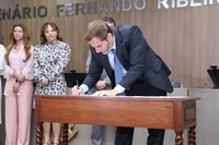 Fotografia em ambiente institucional mostra um homem de terno escuro, camisa azul-clara e gravata azul inclinando-se sobre uma mesa de madeira para assinar um documento com uma caneta preta. Ao fundo, à esquerda, duas mulheres observam em pé: a primeira veste camisa e calça pantalona rosa-claro, e a segunda usa um vestido estampado em preto e branco. Acima delas, na parede de tom amadeirado, lê-se parcialmente a inscrição em letras metálicas "ENÁRIO FERNANDO RIDEI...".