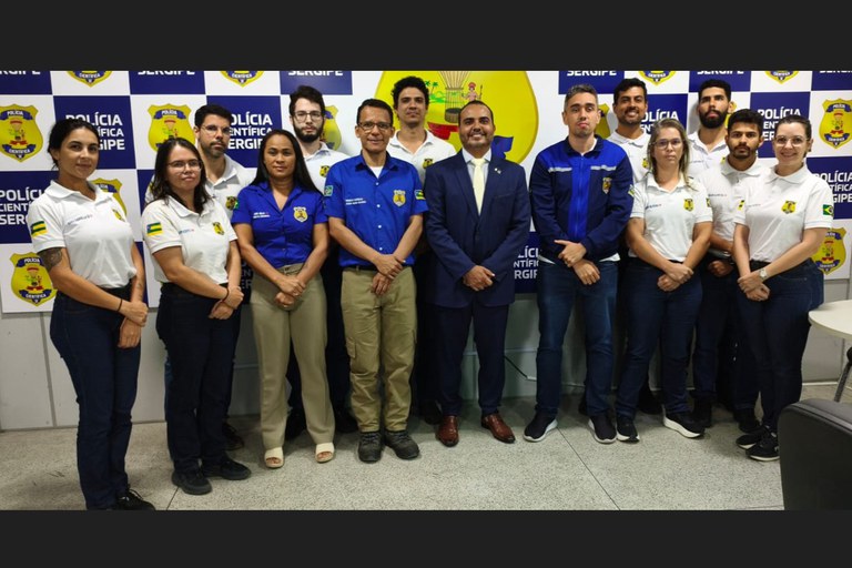 Um grupo de treze pessoas está posicionado lado a lado em pé, sorrindo e olhando para a câmera, em uma sala iluminada. Ao fundo, há um painel azul e branco com o brasão do estado de Sergipe e a inscrição "Polícia Científica Sergipe". A maioria das pessoas veste uniformes institucionais com o brasão no peito ou na manga, variando entre camisas polo brancas, camisas sociais de manga curta azuis e jaqueta esportiva azul. No centro do grupo, destaca-se um homem vestindo terno e gravata azul-marinho. Todos estão em pose formal, com os braços cruzados à frente do corpo ou ao lado.