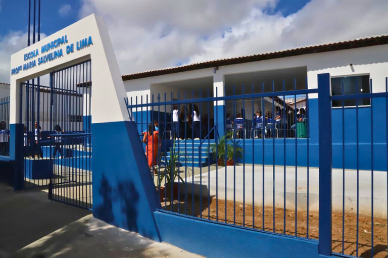 Foto mostra fachada da Escola Municipal Maria Salvelina de Lima, onde se vê o muro pintado de azul e branco e grades azuis; há alunos uniformizados no espaço interno da escola
