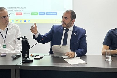 Fotografia em plano médio de um homem de terno azul-marinho, camisa branca e gravata escura, sentado à mesa durante uma reunião. Ele tem barba curta, segura papéis com a mão esquerda e gesticula com a mão direita erguida enquanto fala. Sobre a mesa à sua frente, estão dispostos um microfone de haste, um microfone de mão, uma garrafa térmica preta e folhas de papel. À sua direita na imagem, outro homem de camisa social branca, óculos e crachá o observa atentamente, enquanto ao fundo projeta-se na parede a tela de um sistema digital com botões coloridos de navegação.