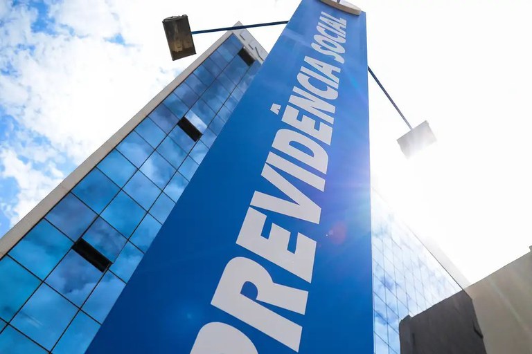 Esta imagem apresenta uma perspectiva de ângulo baixo e dramática de uma unidade da Previdência Social no Brasil, destacando um grande totem vertical azul em primeiro plano com o nome da instituição em letras brancas imponentes. O totem se estende em direção ao céu, ladeado por um edifício moderno de fachada espelhada que reflete o azul celeste e algumas nuvens esparsas. A iluminação solar intensa cria um efeito de brilho direto (lens flare) no canto direito, conferindo uma sensação de magnitude e oficialidade à estrutura arquitetônica.