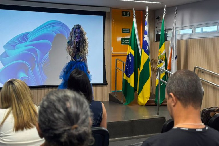 Foto mostra auditório do evento com o publico de frente para o palco com telão e a procuradora Gisele Bleggi falando ao microfone. Ao lado, três bandeiras