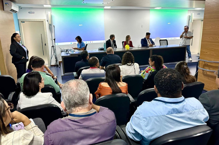 A imagem mostra um auditório com cadeiras pretas ocupadas por diversas pessoas, algumas de costas e outras de perfil. À frente, há uma mesa longa com quatro pessoas sentadas e uma mulher à esquerda, digitando em um notebook. Um homem de camisa clara e jeans fala ao microfone em pé, próximo à mesa. Duas telas de projeção exibem um fundo azul e verde com a sigla ¿MPF¿. No canto esquerdo, uma mulher de terno preto está em pé observando. As paredes são revestidas de madeira e a iluminação é branca. O ambiente sugere um evento formal.