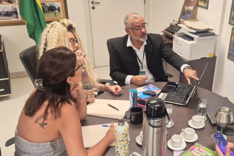 Foto de três pessoas em uma reunião de trabalho em um escritório, com um homem apontando para o laptop e uma mulher de costas anotando em um caderno.