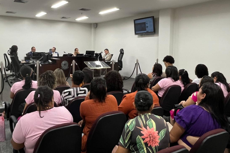 Descrição: Imagem de uma audiência em uma sala de tribunal moderna. No primeiro plano, várias pessoas sentadas em cadeiras pretas, muitas vestindo roupas em tons de rosa, laranja e estampadas. Algumas têm cabelos presos, outras soltos. No centro da sala, há uma bancada elevada com quatro pessoas: duas de terno escuro, uma mulher digitando e um homem de terno claro sentado à parte. Uma tela na parede exibe um conteúdo projetado. A iluminação é branca e forte, com paredes claras e um piso escuro. A sala tem divisórias metálicas e um púlpito de vidro.