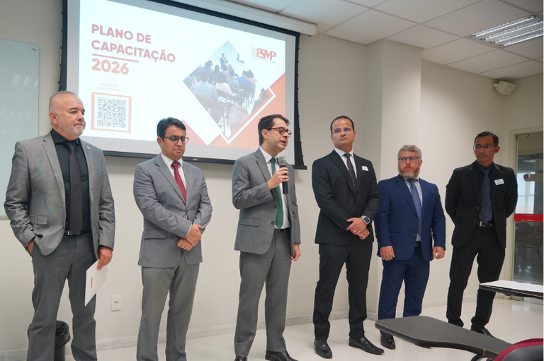 Seis homens vestidos com trajes formais (ternos e paletós) estão em pé, lado a lado, em uma sala de conferências. O homem ao centro fala ao microfone. Ao fundo, uma tela de projeção exibe o título "Plano de Capacitação 2026", o logotipo da ESMP e um QR Code. O ambiente é iluminado e possui paredes claras.