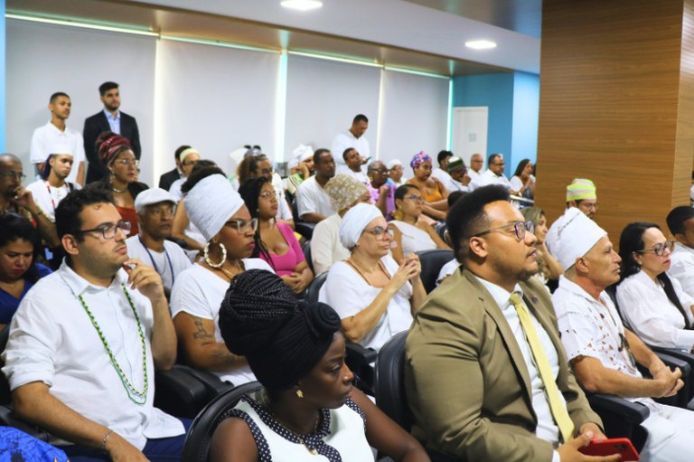 A imagem mostra um grupo de pessoas sentadas em um ambiente interno, possivelmente durante um evento ou reunião. A maioria dos participantes está vestida com roupas brancas ou em tons claros, algumas usando acessórios coloridos, como turbantes e colares. As expressões faciais variam entre concentração e reflexão. No primeiro plano, um homem de terno claro e gravata segura um celular vermelho, enquanto uma mulher ao seu lado, com cabelo trançado, observa atentamente. Ao fundo, há pessoas em pé e sentadas, com destaque para uma mulher com um turbante branco. O ambiente é bem iluminado, com paredes em tons neutros e janelas que permitem a entrada de luz natural.