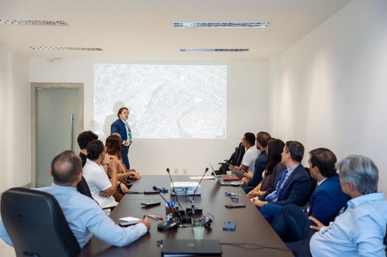foto mostra uma sala de reuniões com onze pessoas entre homens e mulheres usando roupas formais sentadas em uma mesa com microfones, e computadores olhando para a projeção de um mapa na parede enquanto um homem está em pé ao lado