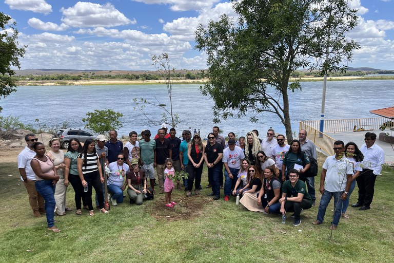 Esta fotografia registra um grupo diversificado de pessoas reunido em uma área gramada às margens de um rio largo, possivelmente o Rio São Francisco, sob um céu aberto com nuvens esparsas. O registro, vinculado ao lançamento de um documentário sobre regeneração ambiental (Borba da Mata), mostra homens, mulheres e crianças de diferentes idades e estilos, sugerindo uma colaboração entre comunidades locais e representantes institucionais (como o MPF e MPSE). No centro da composição, destaca-se uma pequena muda de árvore recém-plantada, simbolizando o esforço coletivo de reflorestamento e preservação da mata ciliar que compõe a paisagem ao fundo.