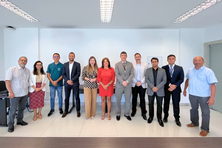 Um grupo de 11 pessoas está posicionado lado a lado em uma sala iluminada, com paredes brancas e piso claro. Todos estão de pé, olhando para a câmera e sorrindo. Há uma diversidade de trajes, incluindo roupas formais, como ternos e vestidos, e roupas casuais, como camisas e calças. Ao fundo, há uma parede lisa com um quadro pequeno e discreto. O ambiente é iluminado por luzes fluorescentes no teto.