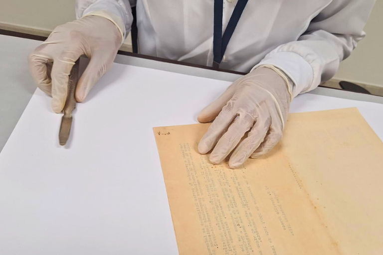 Imagem mostra uma pessoa usando avental branco e luvas de procedimento transparentes, manipulando delicadamente um documento antigo amarelado, apoiado sobre uma superfície branca. Com a mão direita, a pessoa segura uma bisturi, enquanto a mão esquerda mantém o documento esticado. O foco está nas mãos protegidas e no documento, sugerindo um ambiente controlado, possivelmente de restauração ou conservação de papel histórico. A cena transmite cuidado e precisão no manuseio do objeto sob condições que preservam sua integridade.