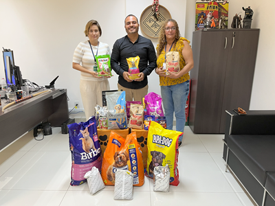 A imagem mostra três pessoas em pé, lado a lado, em uma sala de escritório. Cada uma delas segura um pacote de ração para animais. No chão, à frente do grupo, há vários sacos e pacotes de ração para cães e gatos, de diferentes marcas e tamanhos.