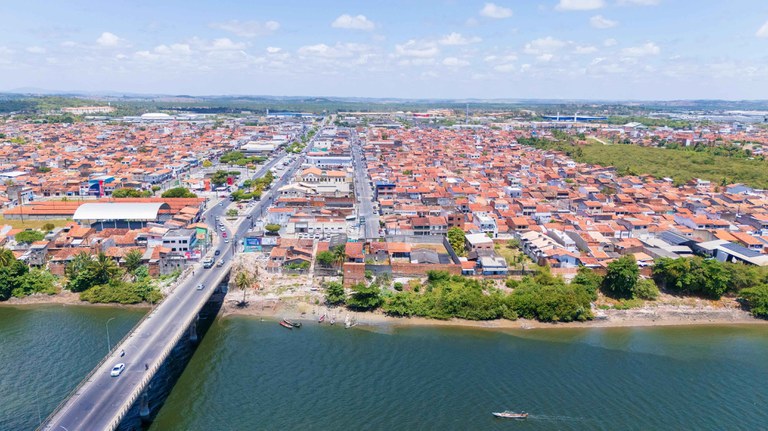 Vista aérea da cidade de Nossa Senhora do Socorro, em Sergipe, destacando a Ponte sobre o Rio do Sal no canto inferior esquerdo. A imagem mostra o contraste entre as águas esverdeadas do rio, a densa área urbana composta por casas de telhados alaranjados e a vegetação de manguezal à direita. O céu está claro e o clima é ensolarado.