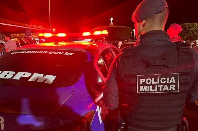 Fotografia noturna em plano médio de costas de um policial militar com colete com a identificação "Polícia Militar".
Ao lado do policial, uma viatura da Polícia Militar de Sergipe com girofleds vermelhos ligados.