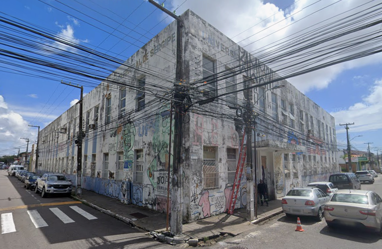 Fotografia de um prédio de dois pavimentos em uma esquina no centro de Aracaju. A fachada está desgastada e coberta por grafites, incluindo a inscrição "João Mulungu". No topo, lê-se "Universidade Federal de Sergipe". Há postes com muitos fios elétricos à frente e carros estacionados na lateral sob um céu azul com nuvens.