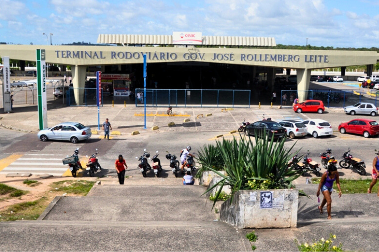 A imagem mostra a entrada do Terminal Rodoviário Governador José Rollemberg Leite. O terminal é um prédio grande, com um teto elevado e a inscrição do nome em letras grandes na parte superior. À frente, há uma faixa de pedestres e um estacionamento com vários carros e motos. Algumas pessoas estão caminhando, enquanto outras estão paradas, possivelmente esperando transporte. Ao fundo, o céu está claro com algumas nuvens. O ambiente parece movimentado, típico de um terminal rodoviário, com pessoas indo e vindo.