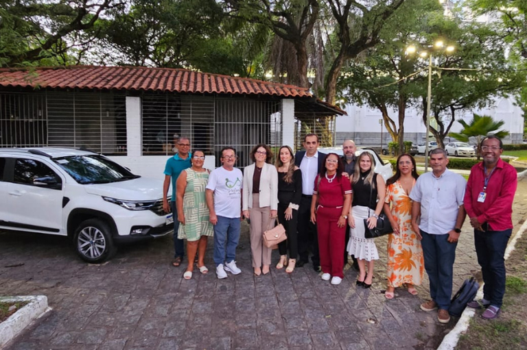 Foto mostra seis homens e seis mulheres em pé, em área aberta, à frente de duas caminhonetes brancas, com uma estrutura em alvenaria atrás, sob árvores
