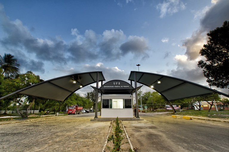 Entrada principal da Universidade Federal de Sergipe, com guarita central e duas coberturas metálicas curvas em destaque sob um céu parcialmente nublado.