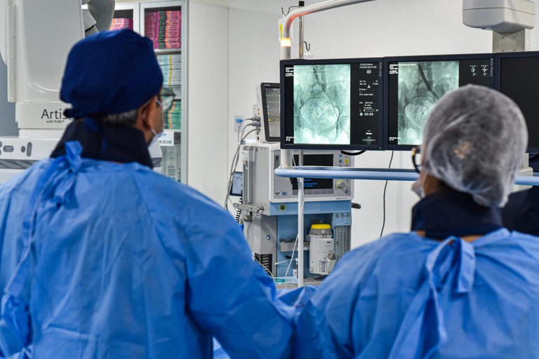 Fotografia em plano médio, capturada de costas, mostrando dois profissionais de saúde em uma sala cirúrgica ou de hemodinâmica altamente equipada. Eles estão observando monitores com exames de imagem em tempo real.
