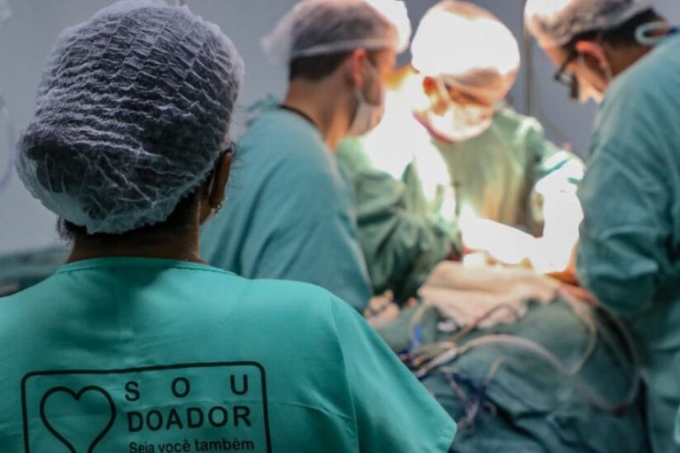 Fotografia colorida em plano detalhe e vista superior de uma cirurgia de transplante de córnea, em um centro cirúrgico. A imagem foca no olho do paciente, coberto por um campo cirúrgico azul que deixa apenas a região ocular exposta. O cirurgião usa luvas azuis e pinças delicadas para manusear a córnea, que é o tecido transparente e brilhante no centro do campo operatório. Ao redor do olho, é possível ver o equipamento e o instrumental cirúrgico, como fios e cânulas finas, indicando a precisão do procedimento oftalmológico. A iluminação é focada e brilhante sobre o olho. Há uma profissional de saúde de costas para a câmera e três médicos realizam o procedimento cirúrgico no fundo da imagem, em uma área desfocada.