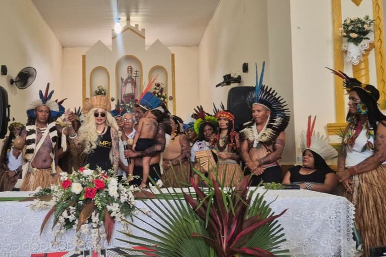 Fotografia colorida de um grupo de indígenas em um ambiente interno; eles usam cocares, pinturas corporais, colares e saias de palha. A mesa é adornada com folhagens e flores