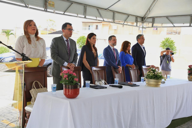 A imagem mostra um evento formal sob uma tenda. Em primeiro plano, há uma mesa coberta com uma toalha branca, decorada com vasos de flores. Atrás da mesa, seis pessoas estão em pé, alinhadas. À esquerda, uma mulher com blusa branca e saia amarela, seguida por um homem de terno cinza e gravata. Depois, uma mulher com vestido preto e um homem com suéter azul. Ao lado, uma mulher de vestido azul e, por fim, um homem de terno escuro. No fundo, um fotógrafo está capturando o momento. O ambiente é iluminado, com vegetação visível ao fundo e veículos estacionados.
