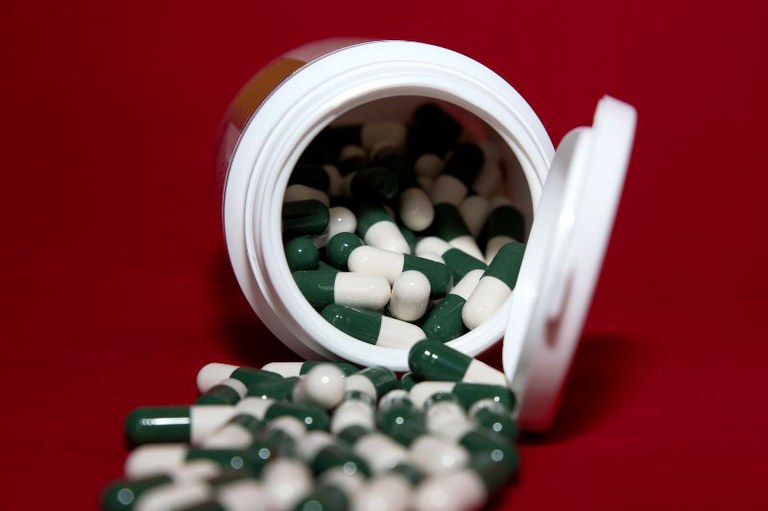 Foto mostra um frasco de remédio aberto em cima de uma mesa e as cápsulas saindo de dentro esparramadas pela mesa vermelha.
