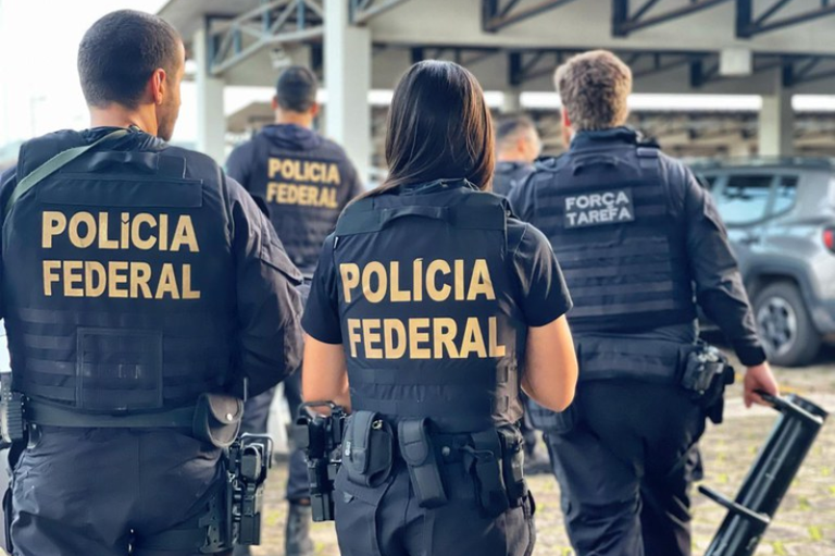 Vista de costas de agentes da Polícia Federal em uma operação. Eles vestem uniformes pretos com coletes balísticos onde se lê "POLÍCIA FEDERAL" em letras amarelas. Uma agente ao centro e outros colegas ao redor aparecem equipados com cintos de utilidades, em um ambiente externo sob uma estrutura coberta.