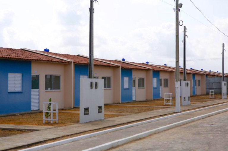 Foto mostra fileira de casas populares geminadas, idênticas, com paredes bege e azul-claro, construídas em um projeto habitacional.