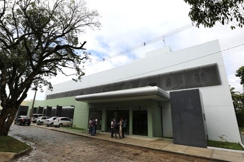 Uma foto em plano médio e ângulo normal mostra a fachada do Hospital Veterinário da UFS, um edifício moderno de dois pavimentos com linhas retas, predominantemente branco e com detalhes em verde-claro na base. No centro, há uma marquise cinza projetada sobre a entrada principal envidraçada, onde um pequeno grupo de pessoas conversa. À esquerda, uma rua de paralelepípedos exibe alguns carros estacionados, parcialmente cobertos pela sombra de uma grande árvore frondosa em primeiro plano. O céu ao fundo está parcialmente nublado, completando o cenário diurno e institucional.