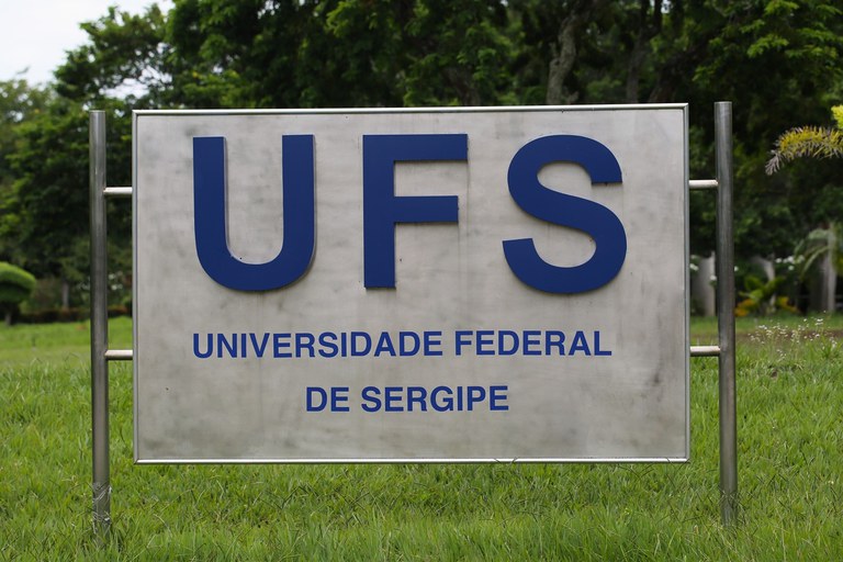 Fotografia de uma placa metálica da Universidade Federal de Sergipe (UFS). Em letras grandes e azuis, no centro da placa, está escrito “UFS”. Abaixo, em tamanho menor, lê-se “Universidade Federal de Sergipe”. A placa está fixada em uma estrutura de metal prateado e posicionada sobre a grama. Ao fundo, há árvores e vegetação.