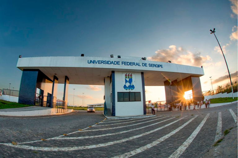 A foto mostra a entrada da Universidade Federal de Sergipe, com uma estrutura ampla e moderna. O portão é de cor branca, com detalhes em azul e o nome da universidade em letras grandes na parte superior. No centro, há um brasão com três chamas. À frente, uma pista de paralelepípedos leva à entrada, onde há um carro branco. Ao fundo, o céu apresenta um tom azul com nuvens e um pôr do sol. Algumas pessoas estão reunidas perto do portão, sugerindo movimento e atividade no local. A iluminação é suave, destacando a arquitetura da entrada.