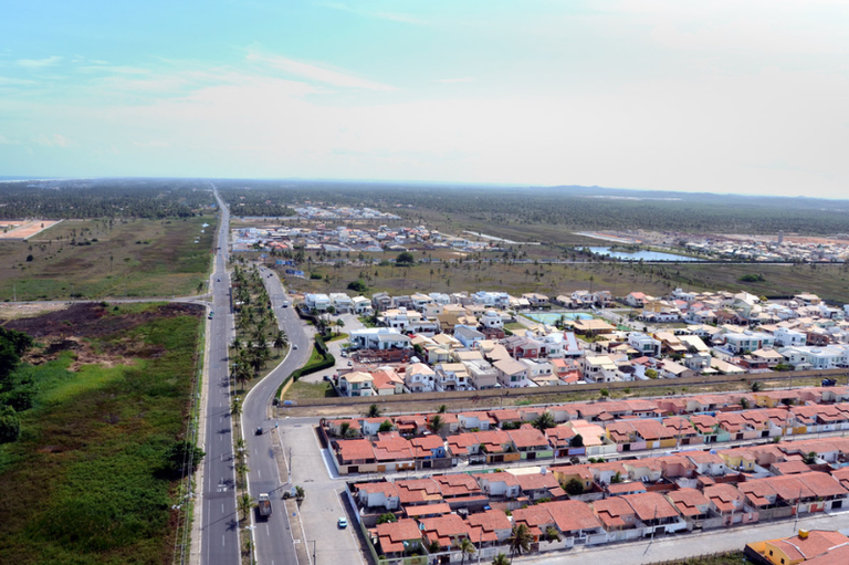 Vista aérea de uma zona de expansão urbana, mostrando um loteamento com casas de telhado cerâmico vermelho organizadas em fileiras. À esquerda, uma longa avenida pavimentada e reta corta a paisagem, ladeada por coqueiros e áreas de vegetação nativa rasteira. Ao fundo, a ocupação urbana se mistura à planície litorânea sob um céu claro.