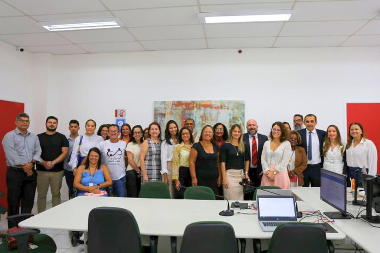 A imagem mostra um grupo de aproximadamente 30 pessoas em um ambiente interno, possivelmente uma sala de reuniões. As pessoas estão dispostas em duas fileiras, algumas sentadas e outras em pé. A maioria está sorrindo e vestindo roupas formais ou semi-formais. Ao fundo, há uma parede branca com uma pintura abstrata colorida e uma parede vermelha. Em primeiro plano, há uma mesa com computadores e equipamentos de apresentação. O ambiente é bem iluminado, com teto branco e piso claro. A diversidade é visível entre os participantes, que incluem homens e mulheres de diferentes idades.
