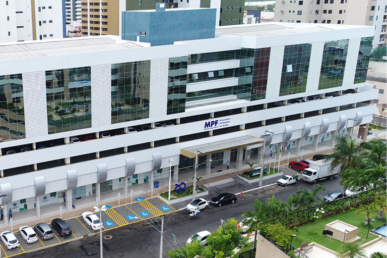 Vista aérea do prédio da Procuradoria da República em Sergipe (MPF). O edifício moderno de cor branca possui janelas espelhadas e a identificação do MPF na fachada, com uma rua e vagas de estacionamento à frente.