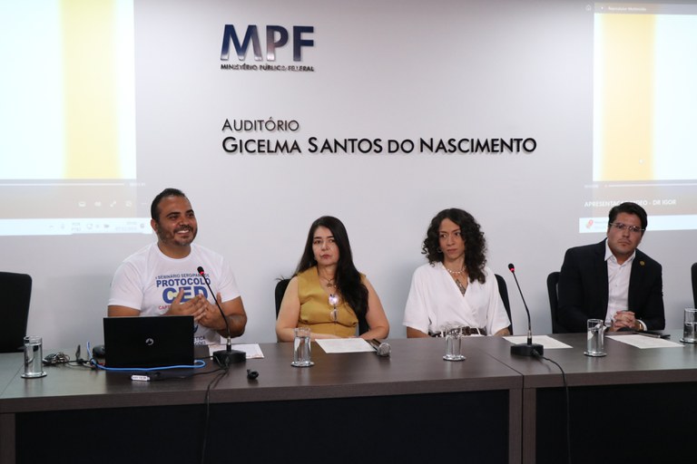 Quatro pessoas estão sentadas lado a lado em uma mesa de auditório do MPF. À esquerda, um homem fala ao microfone diante de um laptop. Ao centro, duas mulheres estão sentadas, e à direita há um homem de terno. Ao fundo, aparece a parede com o nome “Auditório Gicelma Santos do Nascimento”.