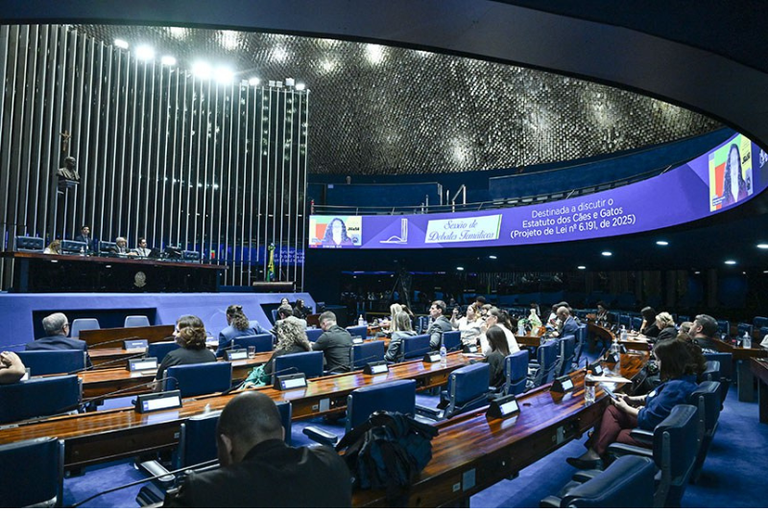 Plenário do Senado com pessoas sentadas em bancadas azuis diante de um grande painel eletrônico curvo que anuncia uma sessão de debates temáticos.