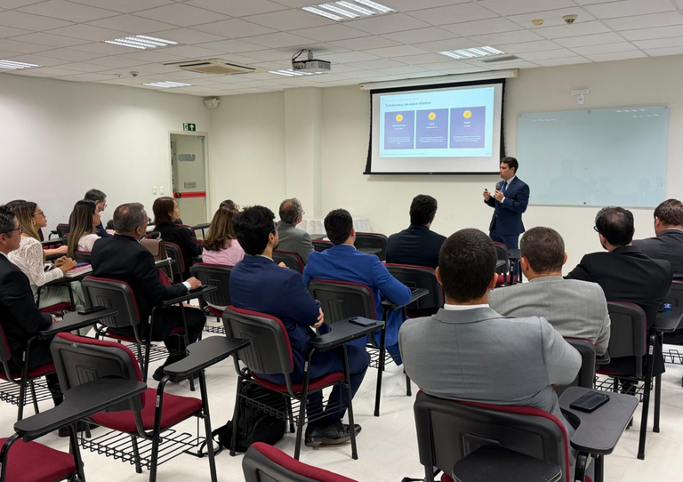 Um homem faz uma apresentação em uma sala de auditório para uma plateia em trajes formais. À direita, o palestrante de pele clara veste terno azul-marinho e gesticula enquanto fala. Ao fundo, uma tela projeta um slide com o título "Estratégias para nova gestão" sobre três colunas roxas com ícones amarelos. Ao lado da tela, há uma lousa branca. Em primeiro plano, diversos espectadores aparecem de costas, sentados em cadeiras estofadas na cor vinho. O ambiente é iluminado, com piso claro e teto modular.