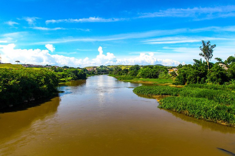 #PraTodosVerem: vista panorâmica do rio Paraíba do Sul sob um céu azul vibrante com nuvens brancas; as águas calmas e acastanhadas são ladeadas por densa vegetação verde em ambas as margens, com colinas suaves e construções urbanas ao fundo.
