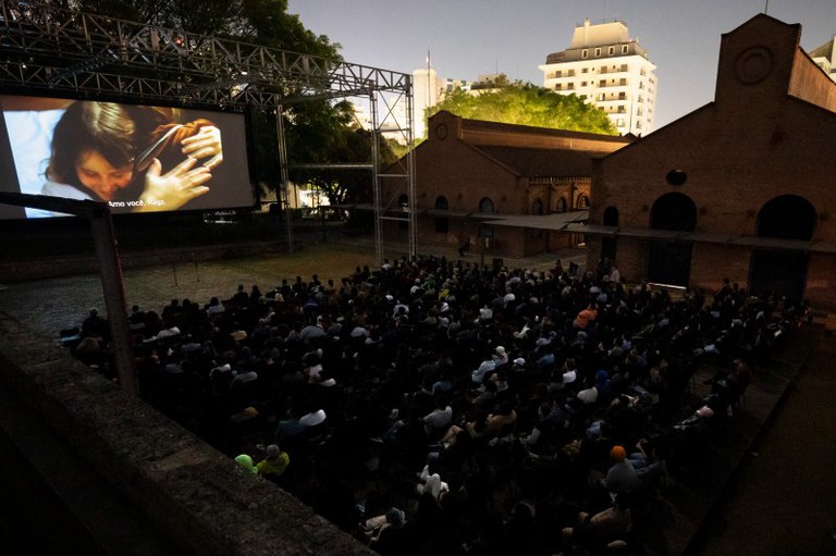 Arte mostra a tela externa da Cinemateca exibindo um filme. A tela é montada na frente do prédio, que tem uma fachada de tijolinhos marrons aparentes