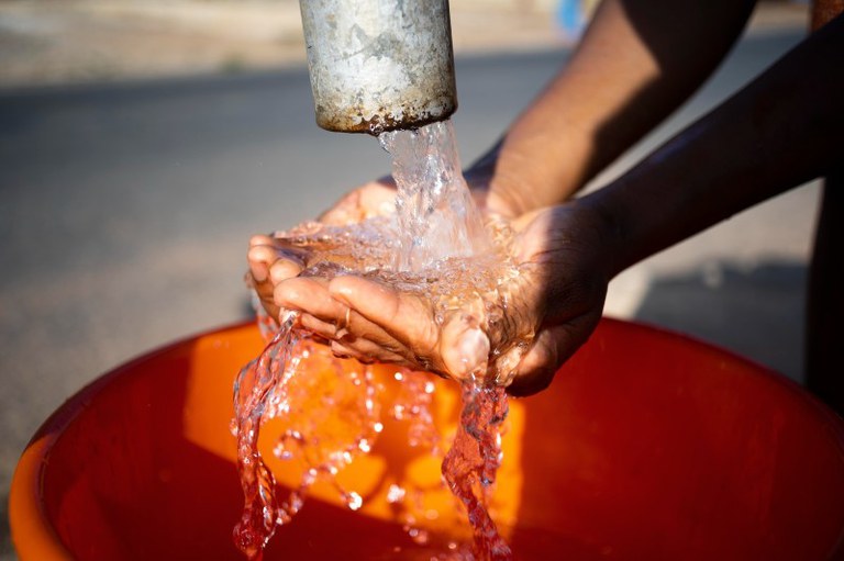 Foto mostra detalhe de um cano de ferro jorrando água enquanto um mão ampara a água que cai em um balde laranja