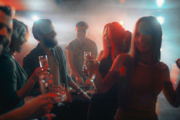 Imagem mostra pessoas em uma festa, com taças na mão e um DJ ao fundo