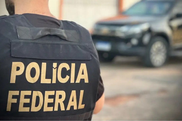 Imagem mostra agente da Polícia Federal de costas, com colete onde se lê o nome da instituição. Ao fundo, uma viatura da PF desfocada