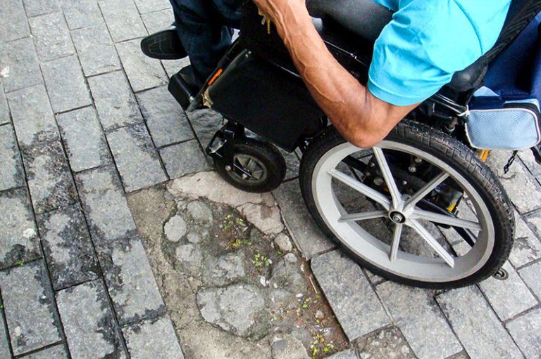Imagem mostra pessoa sentada em cadeira de rodas em uma calçada com falha no revestimento de granito