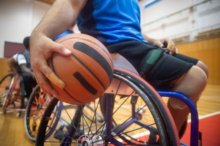A imagem mostra, em primeiro plano e de forma aproximada, um atleta sentado em uma cadeira de rodas esportiva, segurando uma bola de basquete. A foto foi tirada de um ângulo baixo, focando da cintura para baixo do atleta e em sua mão direita, o que destaca a ação e o equipamento.