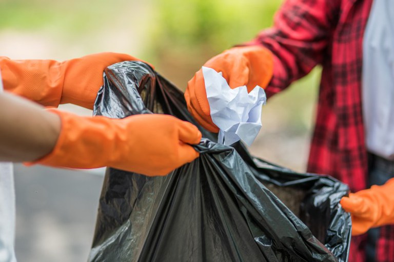 Foto mostra duas pessoas segurando um saco de lixo e jogando papel dentro