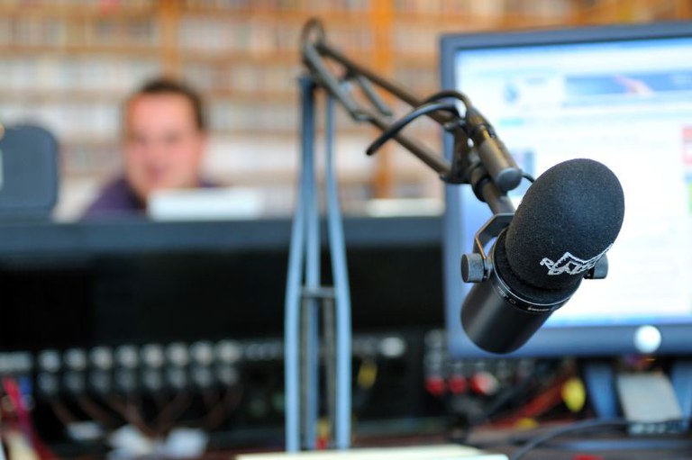 Imagem mostra microfone em estúdio de rádio, ao fundo, um técnico de som aparece sentado, em plano desfocado
