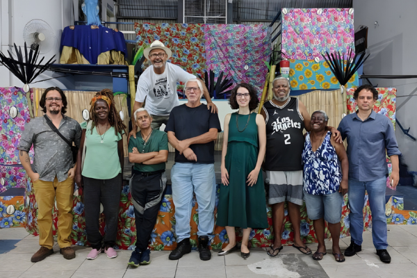 Grupo de pessoas brancas e negras, sendo seis homens e três mulheres. Elas estão em pé, lado a lado. Atrás está um cenário coberto de tecidos estampados
